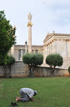 Μουσουλμάνος μετανάστης προσεύχεται εμπρός στην Ακαδημία Αθηνών, πίσω το άγαλμα του θεού Απόλλωνα.